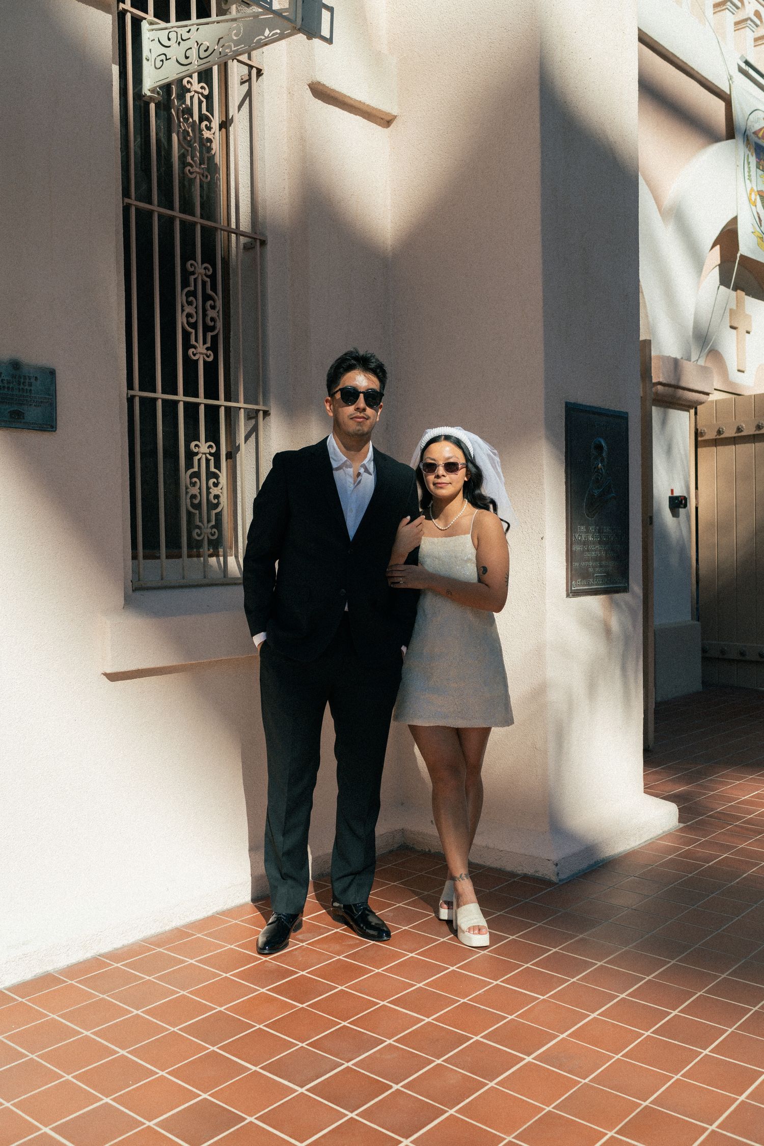 Bride in mini dress and groom in black suit wearing sunglasses posing outside a sunlit building.
