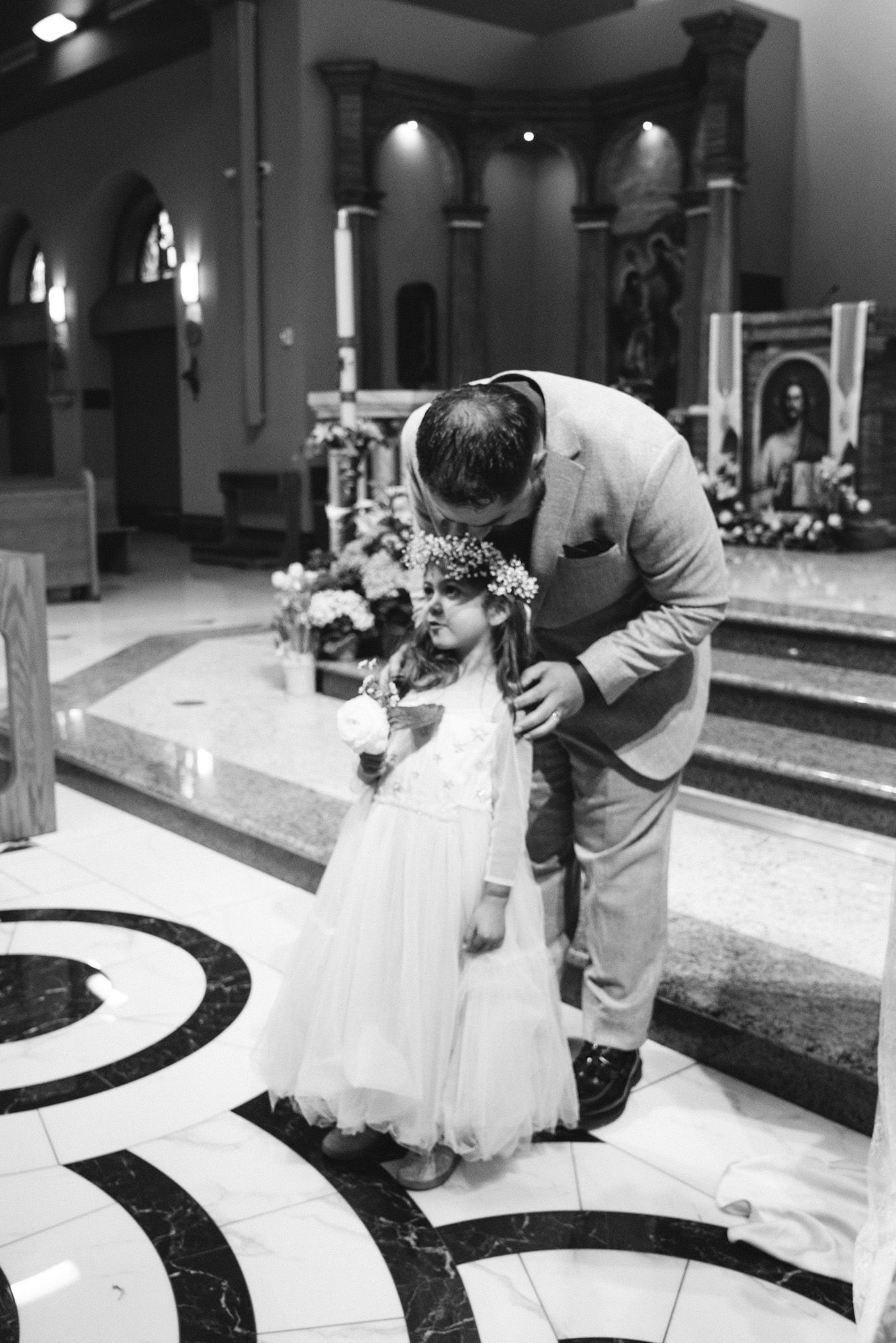 Groom bending down to adjust daughter's flower crown in church after ceremony.