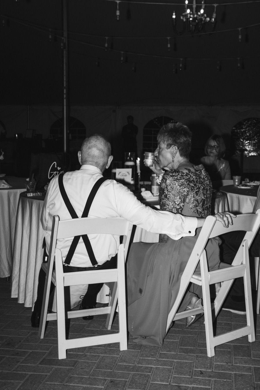 Grandpa with suspenders and grandma in gown share drink at candlelit wedding reception table.