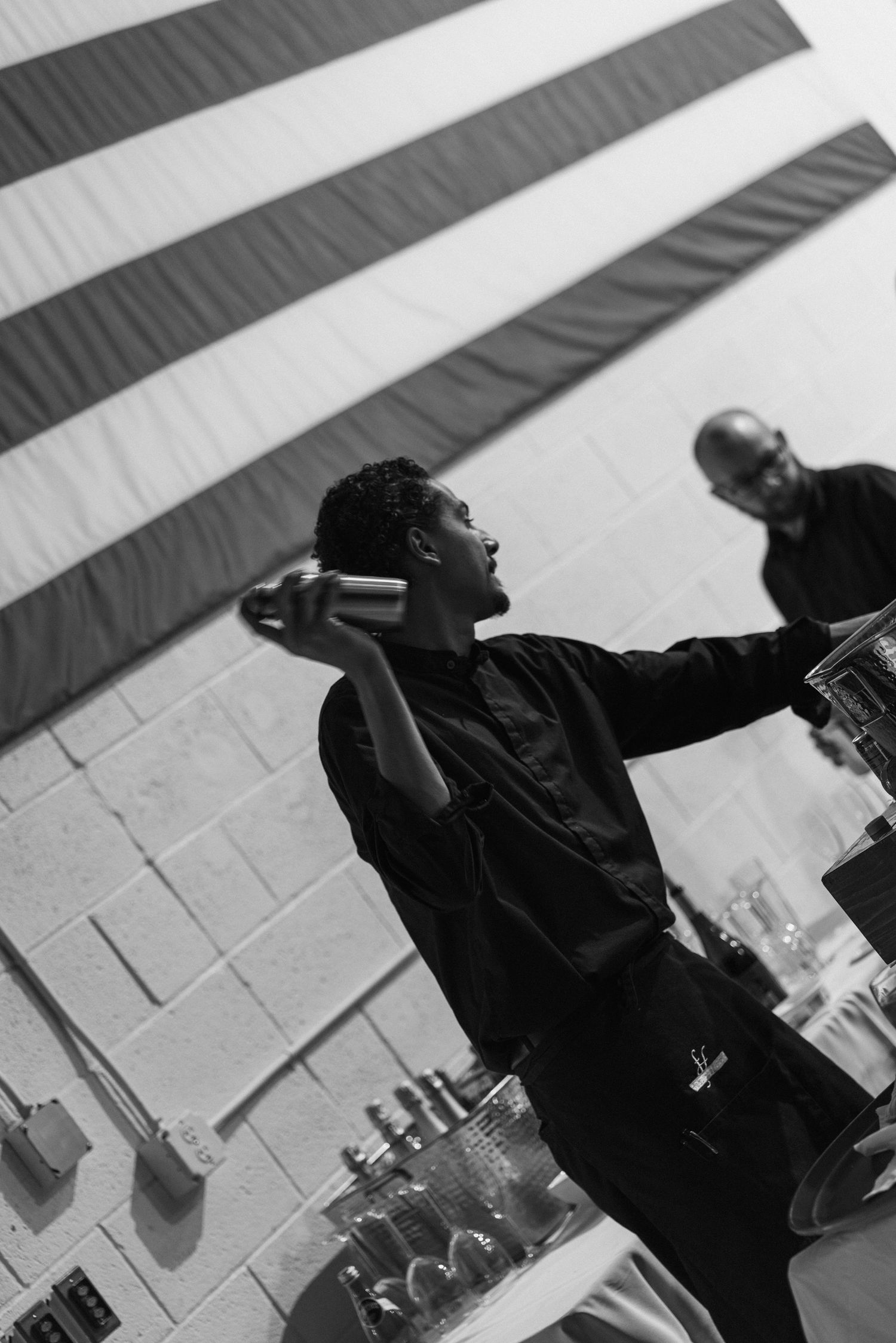 Black and white photo of bartender shaking cocktail shaker beneath large flag at wedding.