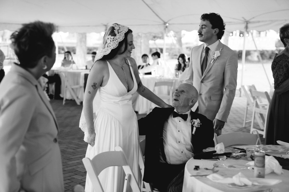 Bride and groom greeting seated elderly guest under wedding reception tent with the mother of the bride nearby, black and white photo.