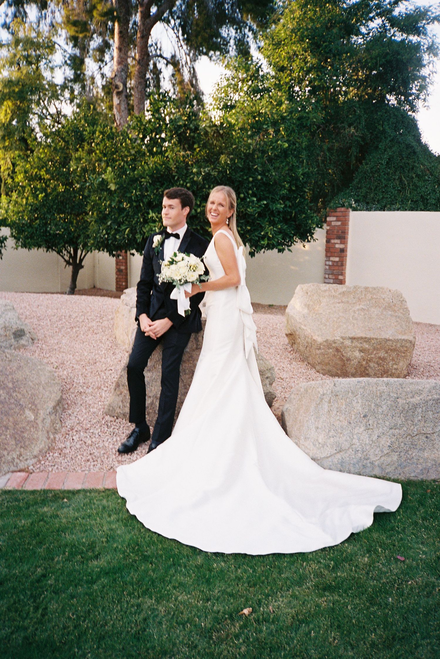 Bride in elegant white gown with train and groom in black tuxedo posing near large desert boulders.