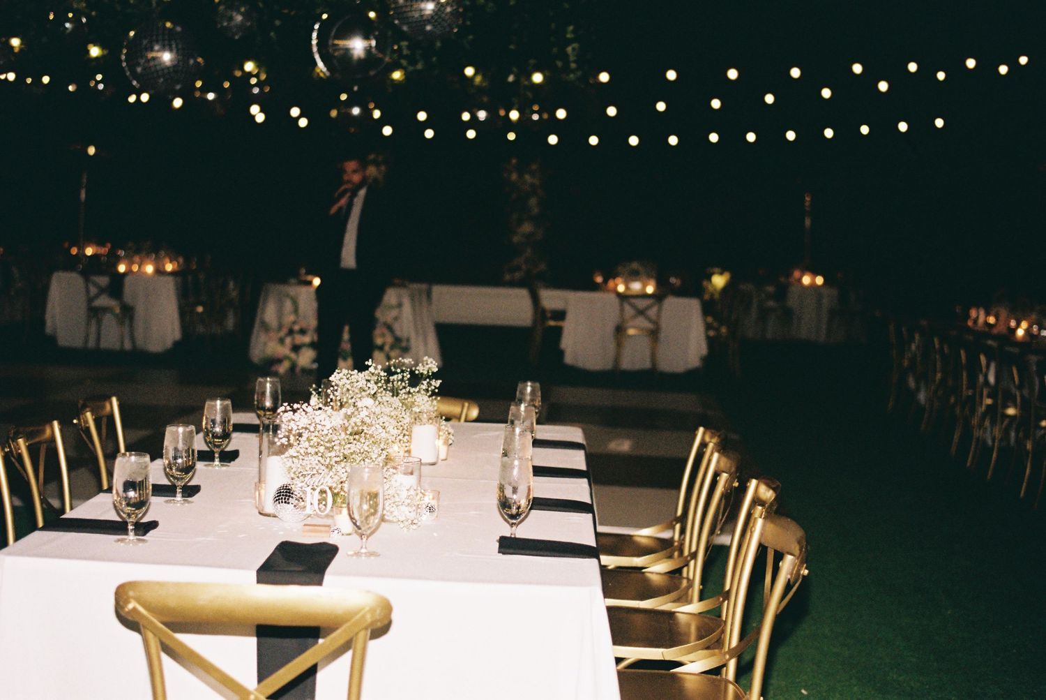 Elegant outdoor wedding reception at night with string lights, white tablecloths, gold chairs, and candle centerpieces.