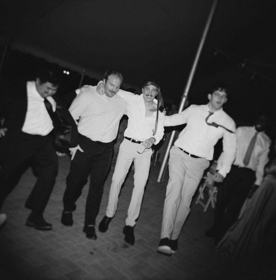 Black and white film photo of groomsmen dancing and jumping together with groom at wedding reception.