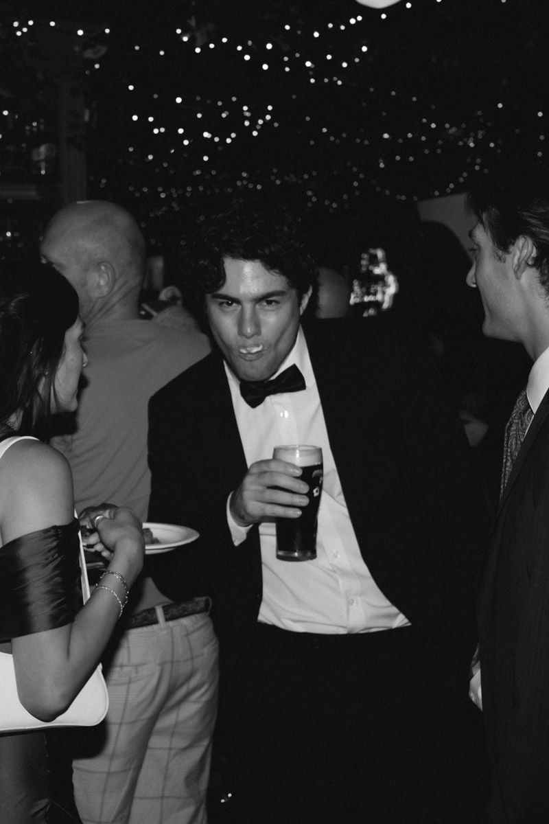 Man in tuxedo holding a pint glass at a formal evening event, black and white photography.