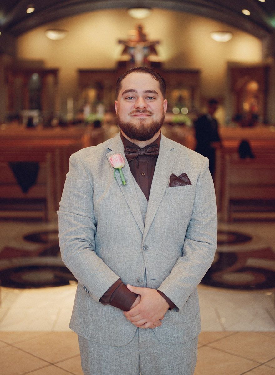 Groom in light gray suit with pink boutonniere and brown bow tie stands in church before wedding ceremony.