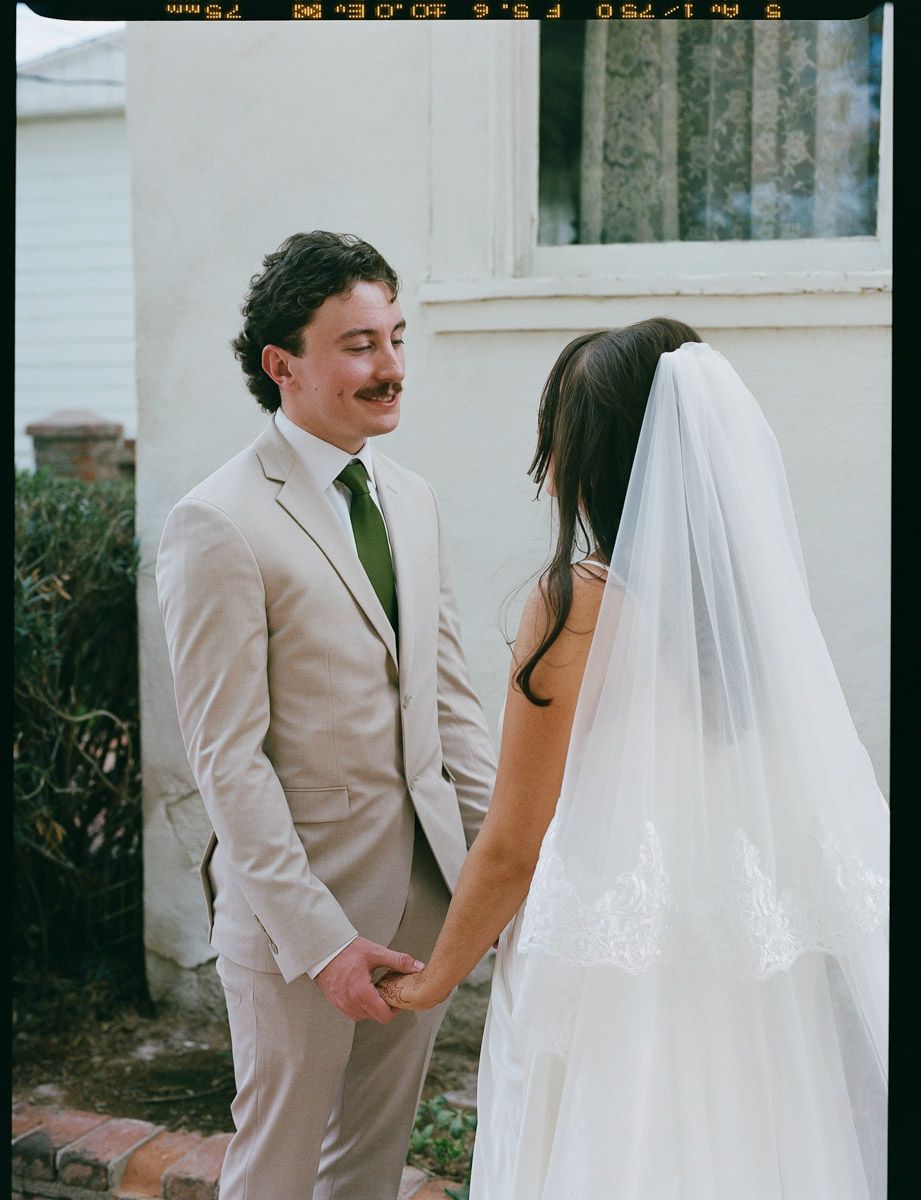A groom in a tan suit holds hands with his bride in a white gown and veil outdoors by a house in Arizona.