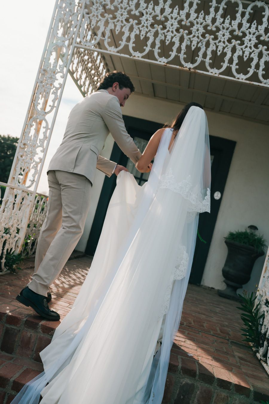 Groom in tan suit helps bride with long veil ascend steps of white building entrance.