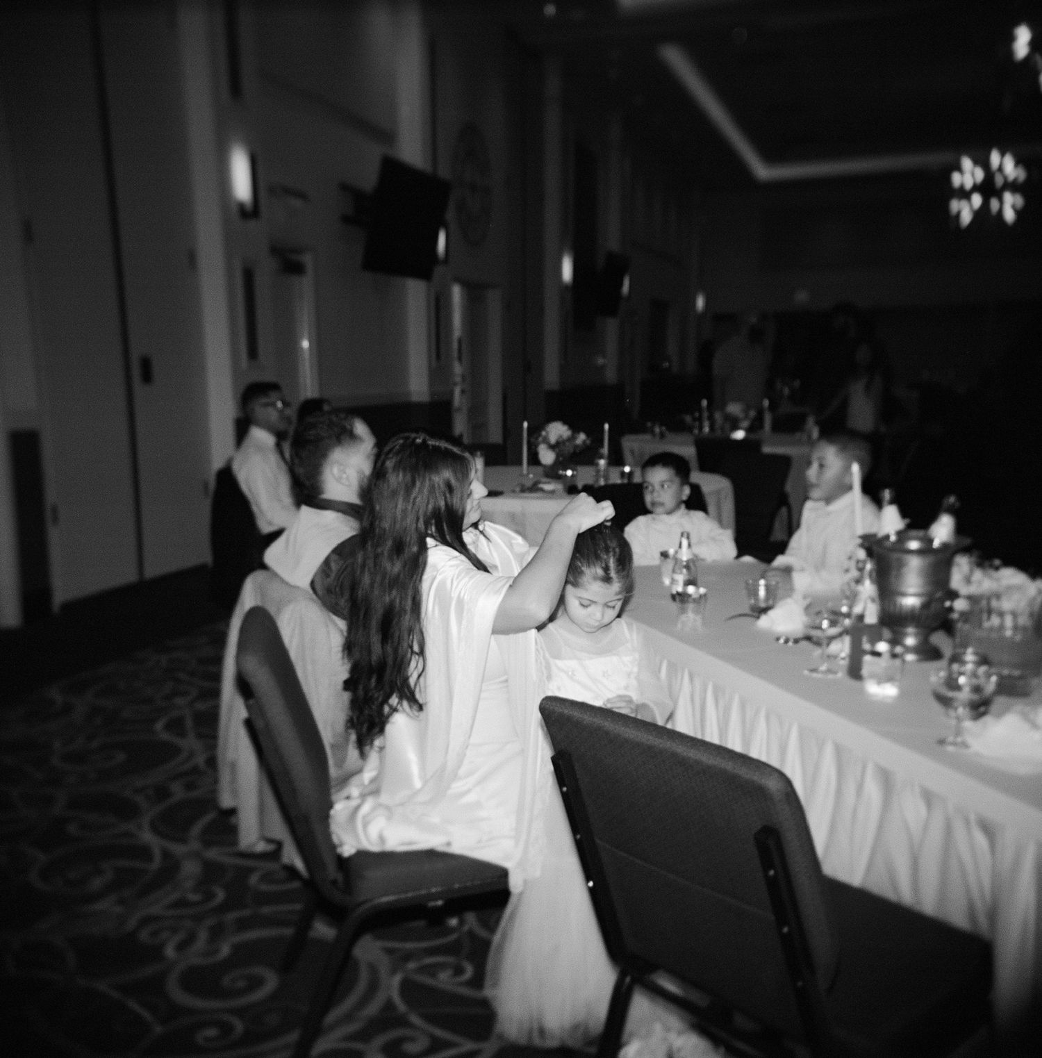 Bride fixing child's hair seated at a wedding reception banquet table in a dimly lit ballroom.