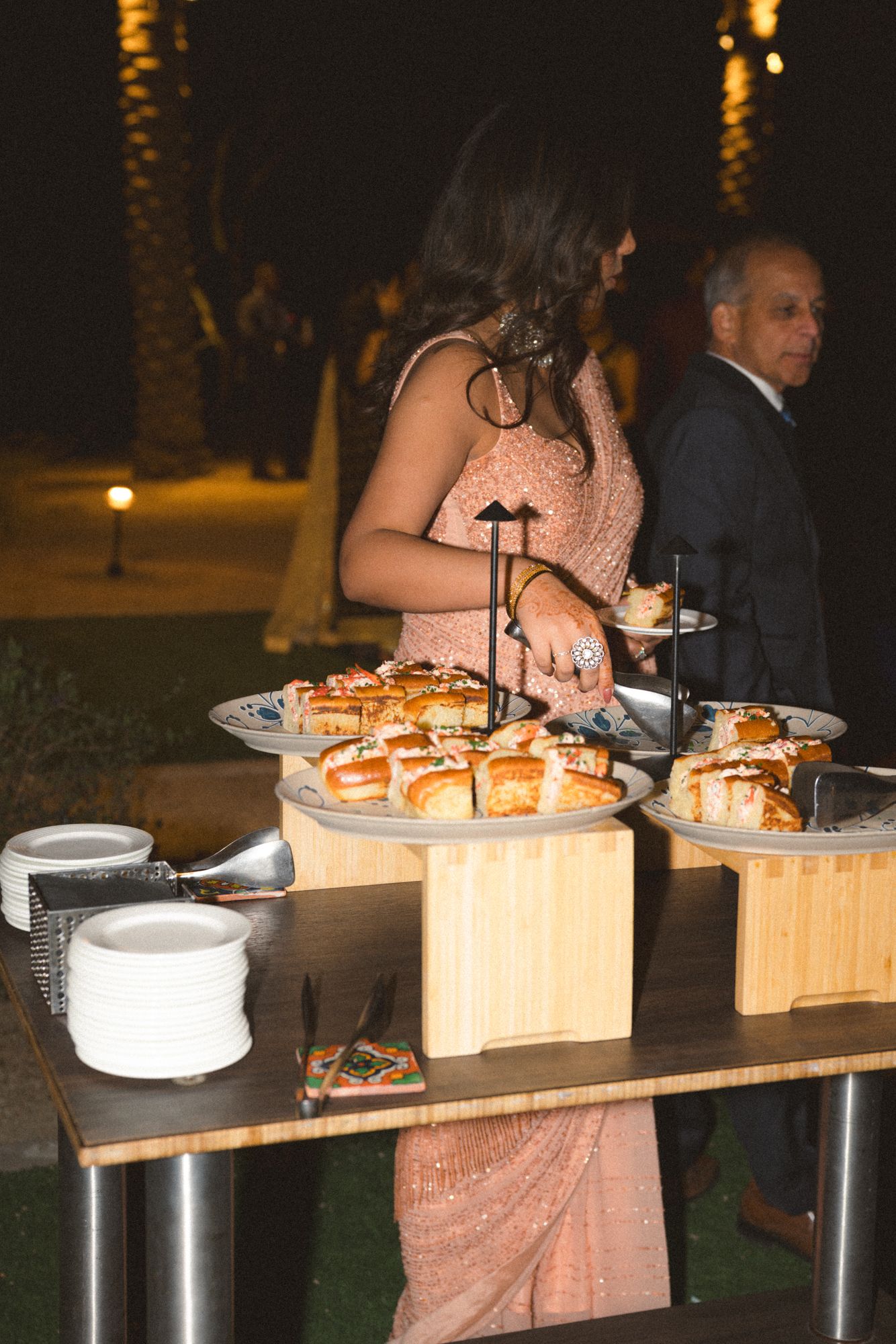 Wedding guest in a pink sequined saree serves herself at an elegant outdoor nighttime food station.