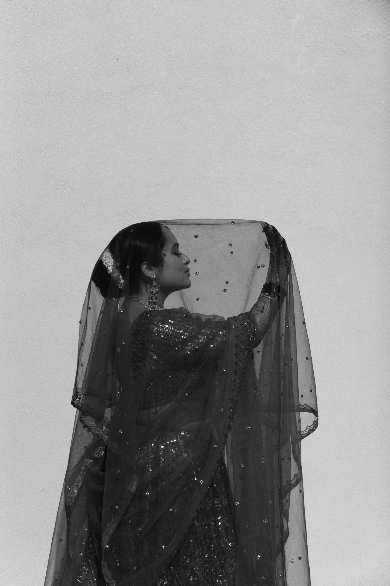 Black and white artistic portrait of a bride in a sequined saree holding a sheer embellished dupatta overhead.