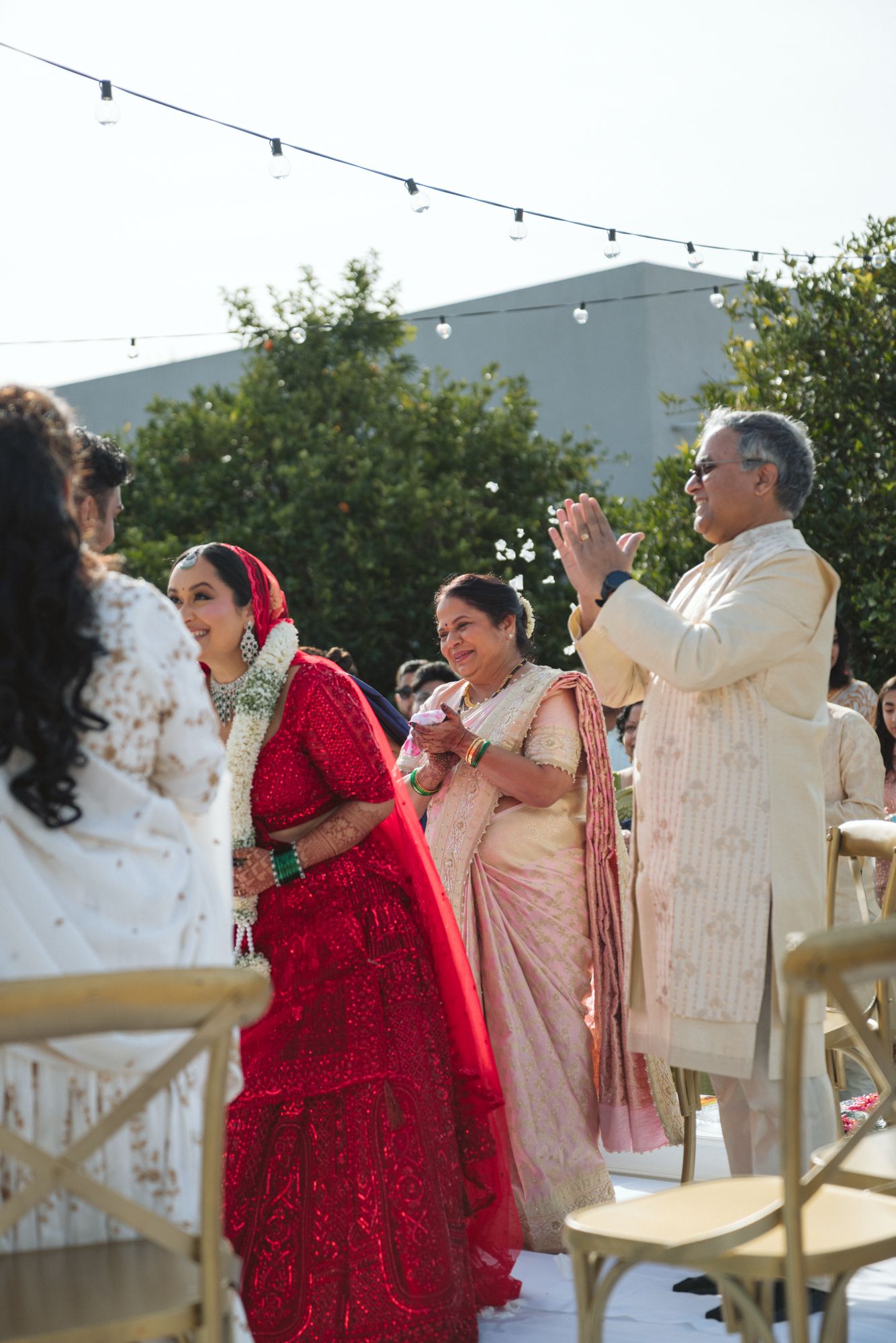 Indian bride in red bridal lehenga choli by her parents at an outdoor ceremony facing the groom.