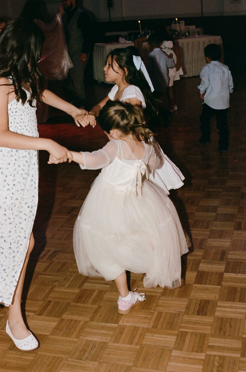Two flower girls in white tulle dresses dance joyfully on a wooden dance floor at a wedding reception.