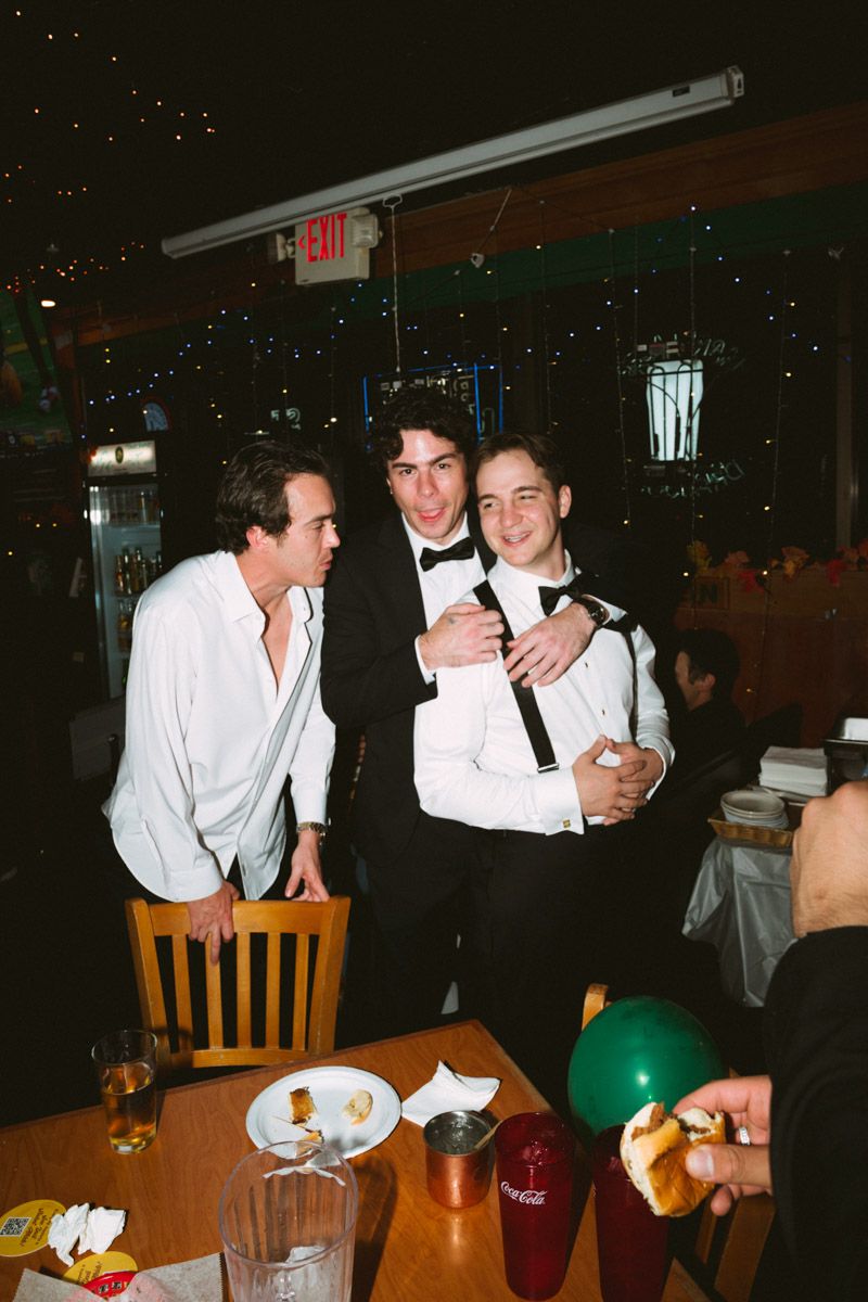 Three young men in formal attire smile together at a restaurant table with drinks and a green balloon.