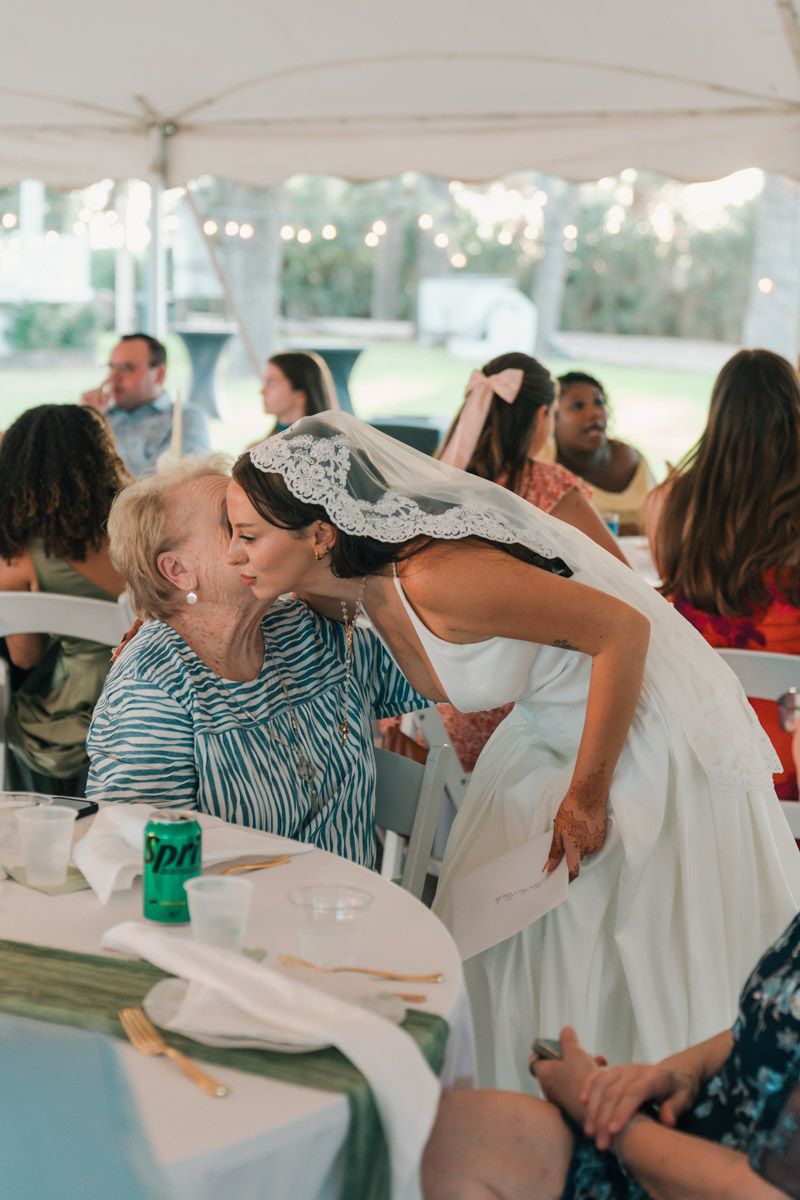 Bride in white gown and lace veil leaning down to kiss elderly guest at outdoor wedding reception under tent.