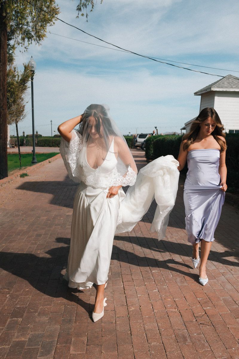 Bride in white lace gown and veil walks with maid of honor in lavender dress on brick path outdoors.