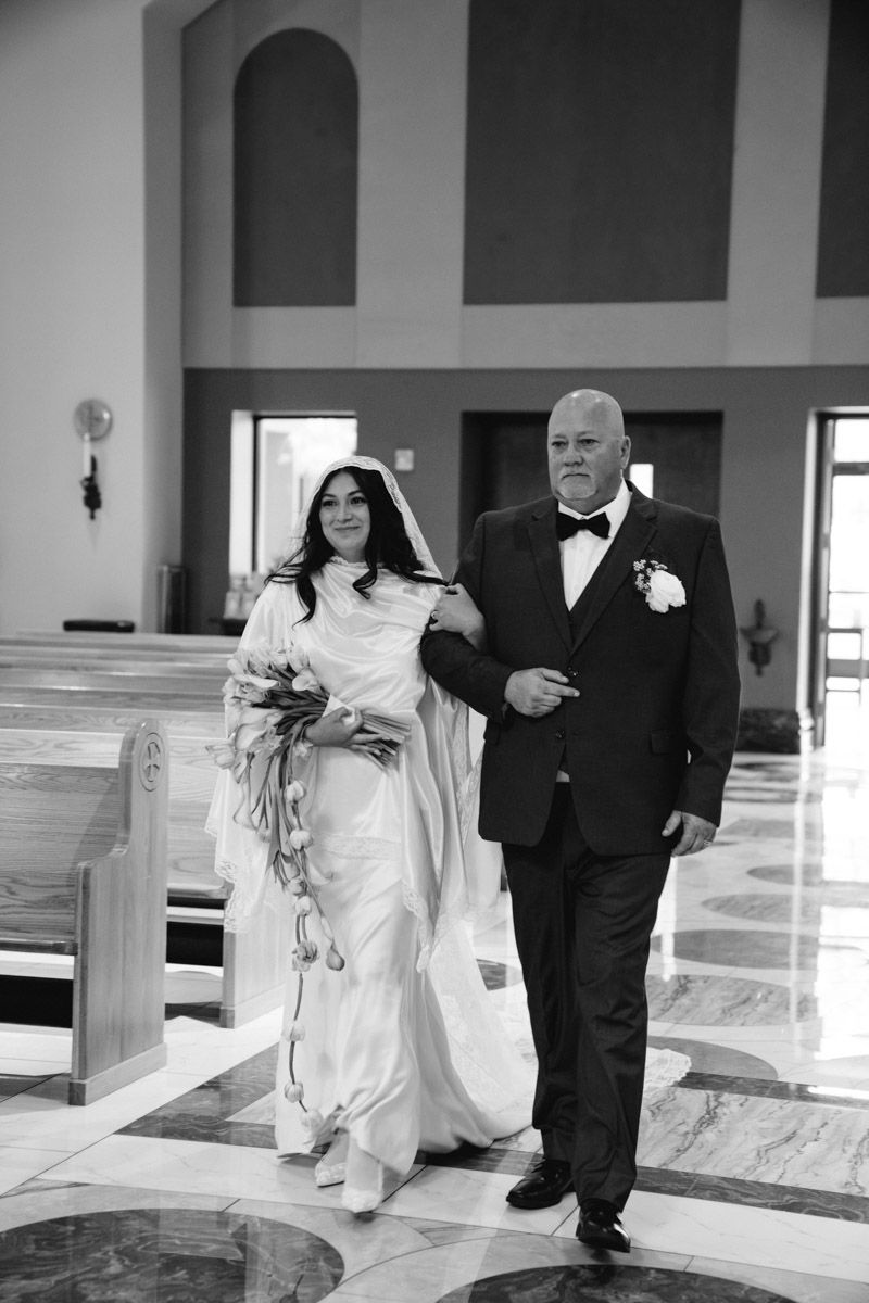 Bride in white gown and veil walks down church aisle with her father in tuxedo in black and white photo.