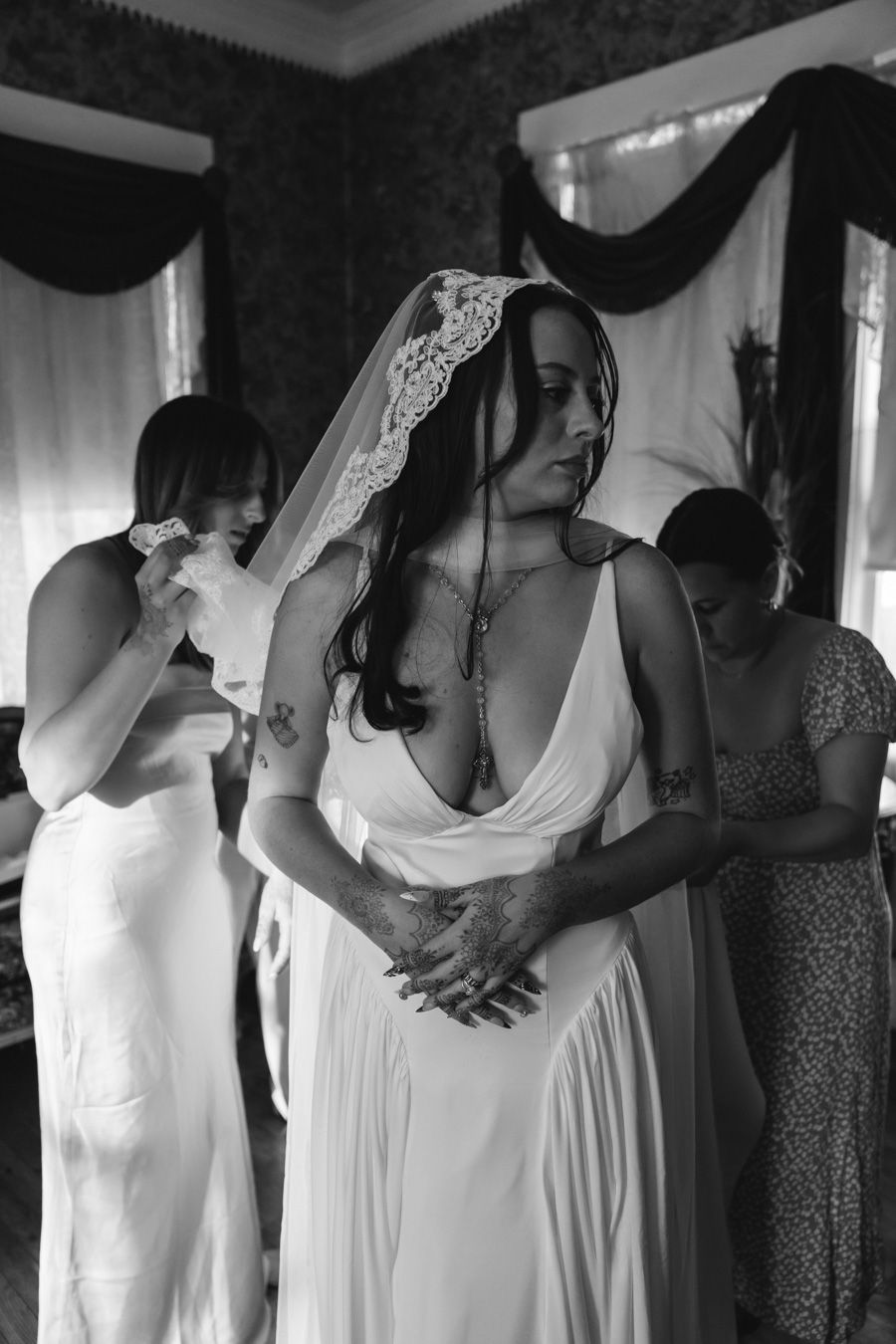 Bride in white gown with lace veil stands with hands clasped as bridesmaids assist in black and white photo.