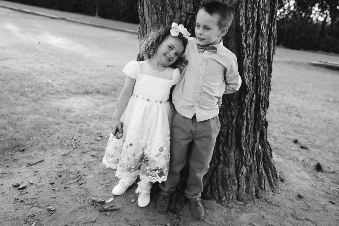 Black and white photo of young flower girl and ring bearer leaning against tree trunk.
