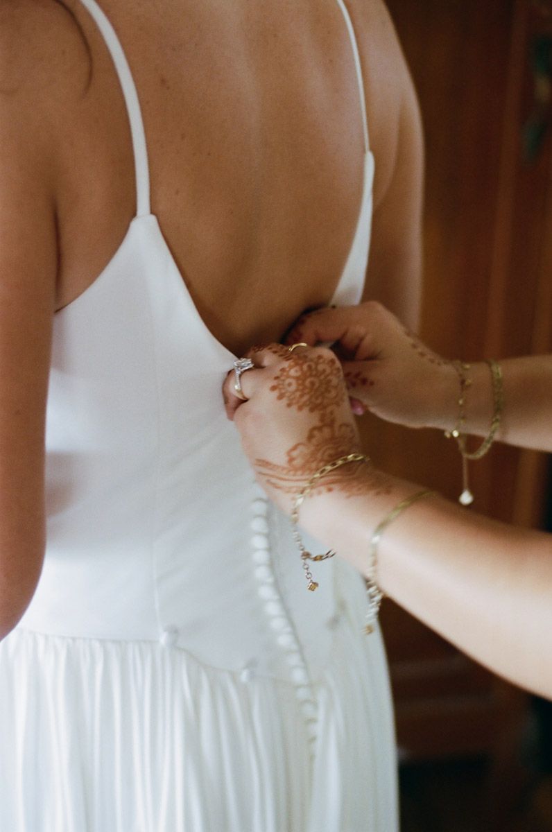Bridesmaid fastening buttons on bride's white wedding dress, hands adorned with henna and gold jewelry.