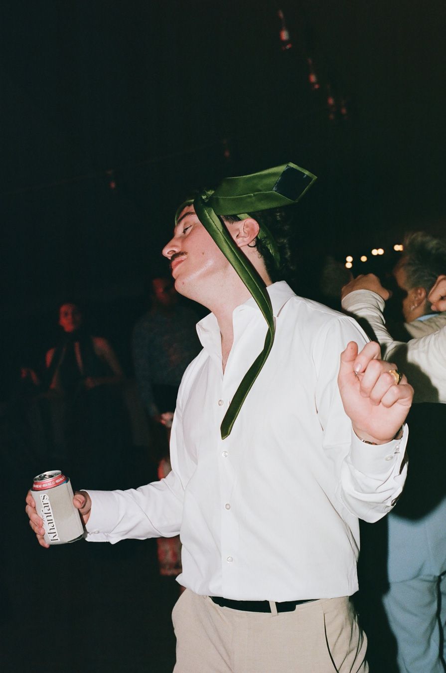 Groom dancing joyfully at wedding reception wearing green tie on head while holding beer can.