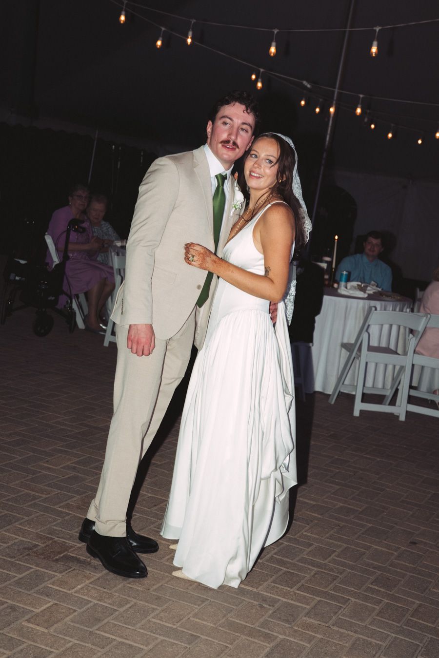 Smiling bride and groom pose together on dance floor under string lights at evening wedding reception.