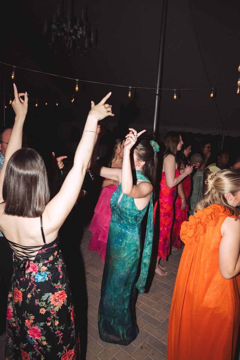 Guests in colorful formal gowns dancing at an outdoor wedding reception under string lights at night.