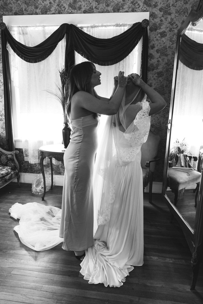 Black and white photo of the maid of honor helping bride with wedding dress in elegant vintage room.