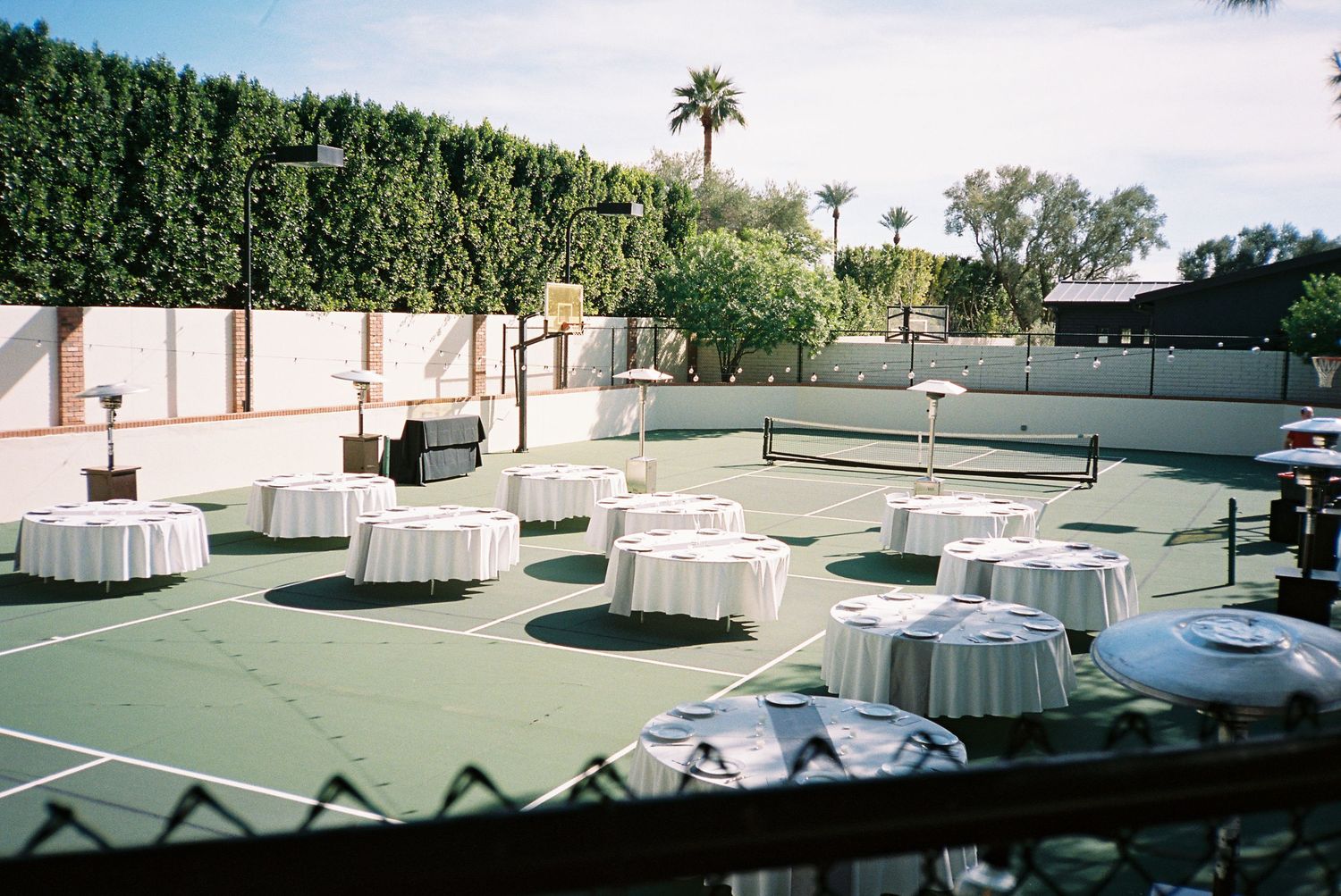 Outdoor wedding reception setup on tennis court with round white-draped tables and greenery.
