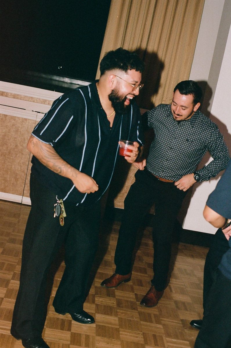 Two men laughing and dancing together at an indoor party, one holding a red drink.