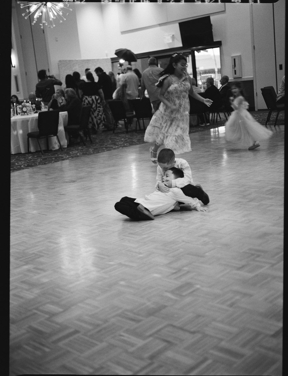 Black and white film photo of a child sliding on a dance floor at a wedding with other kids dancing in the background.