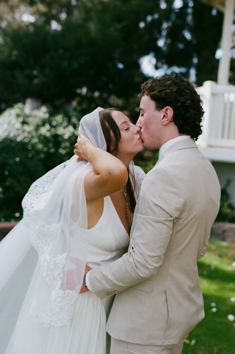A bride and groom share a romantic kiss outdoors, she in a lace veil and white gown, he in a tan suit.