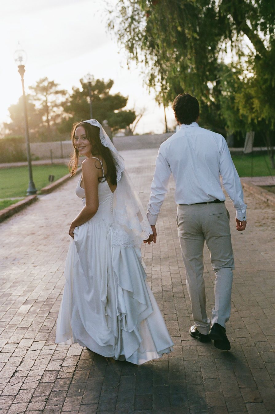 Bride in a flowing white gown and veil walks hand-in-hand with groom at golden hour outdoors down a brick road.