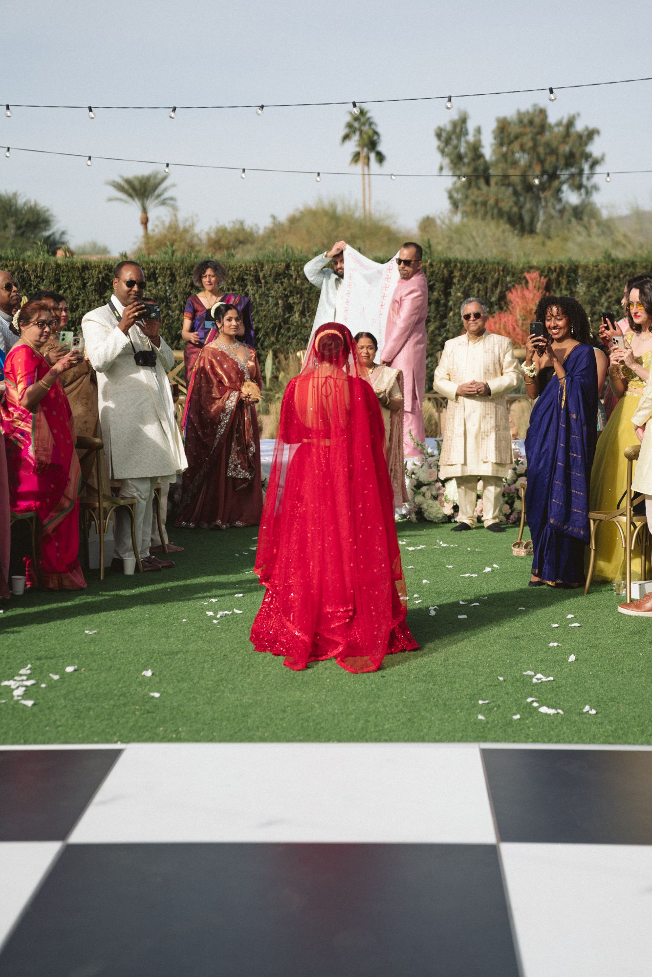 Indian bride in red lehenga choli walks down the aisle at an outdoor wedding ceremony with guests applauding.