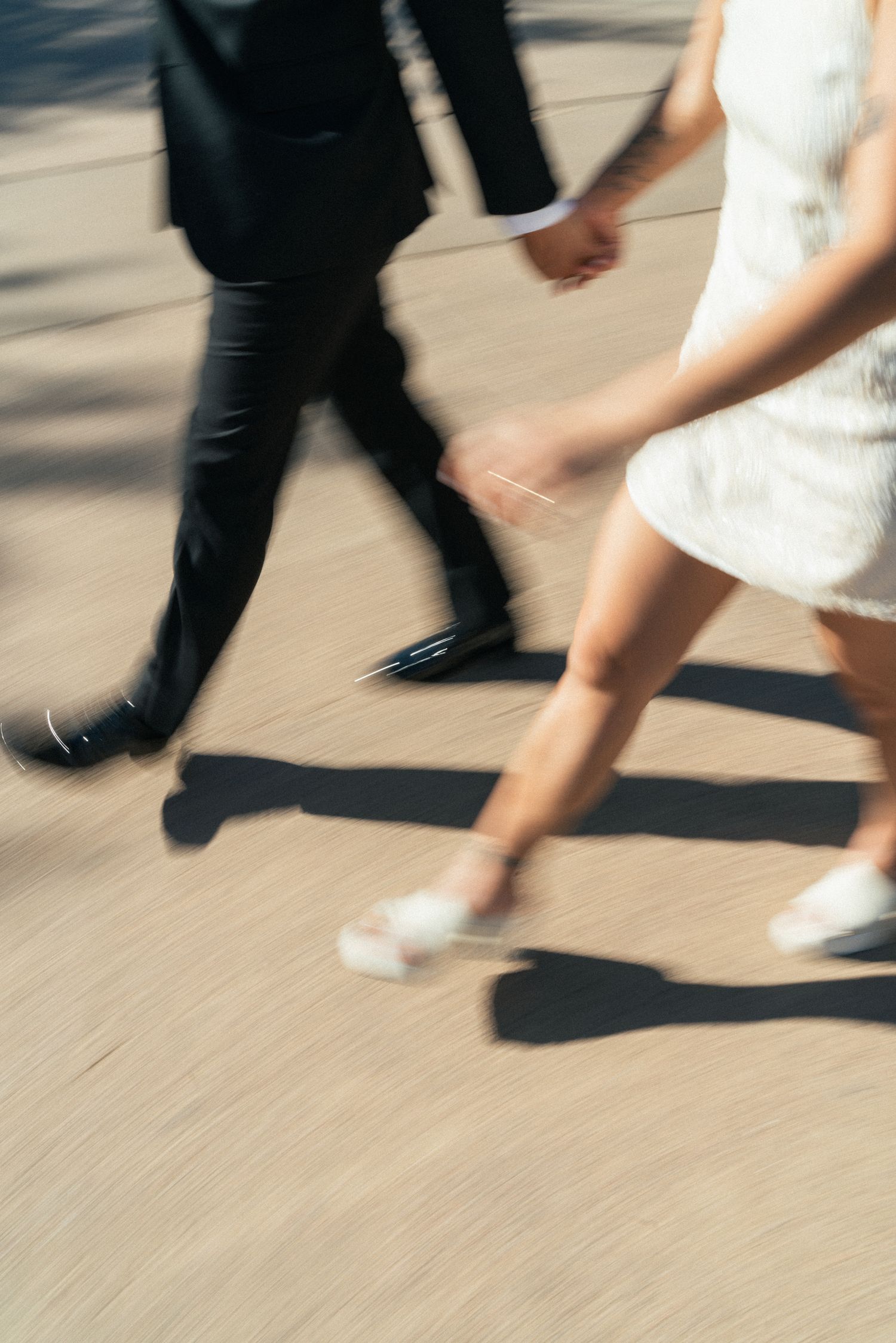 Motion blur photo of bride and groom walking hand in hand on sunny pavement.
