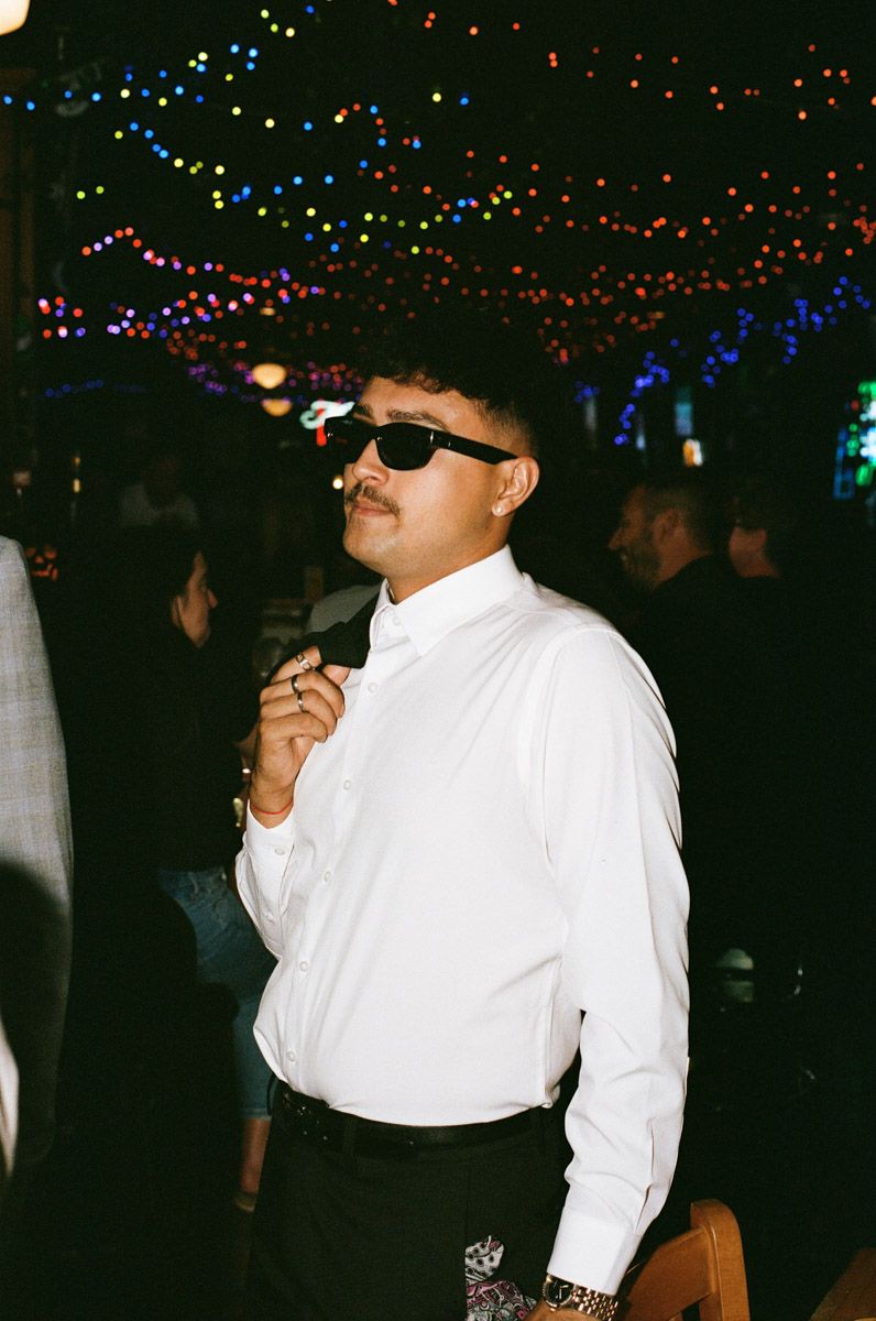 Man in white shirt and sunglasses poses at a colorful night event with bokeh string lights in background.