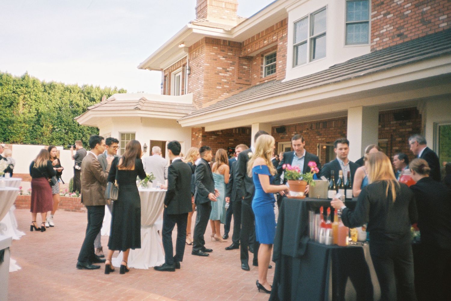 Elegantly dressed wedding guests socializing outdoors near a brick building during a cocktail hour reception.