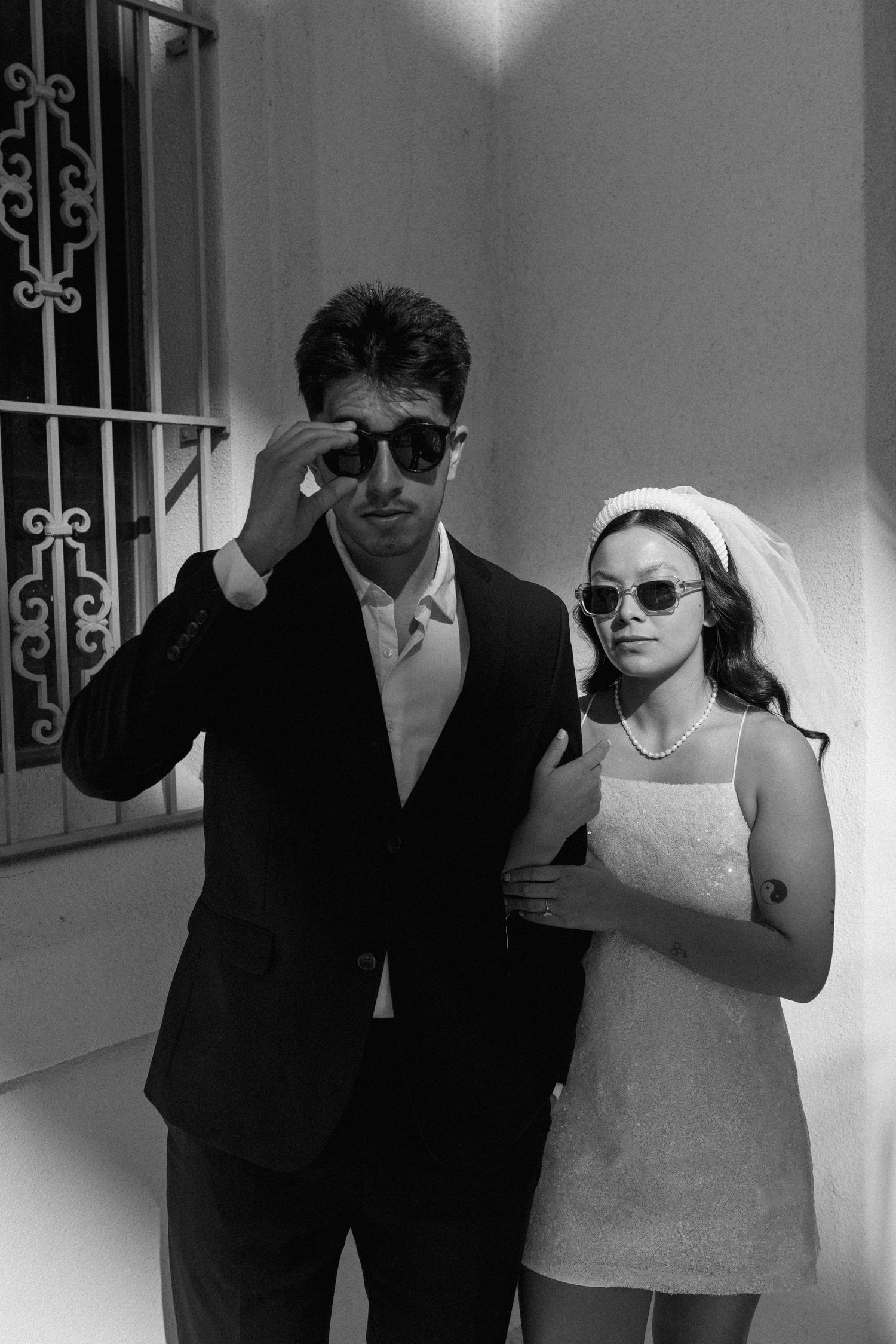 Black and white photo of stylish bride and groom wearing sunglasses posing outside a building.