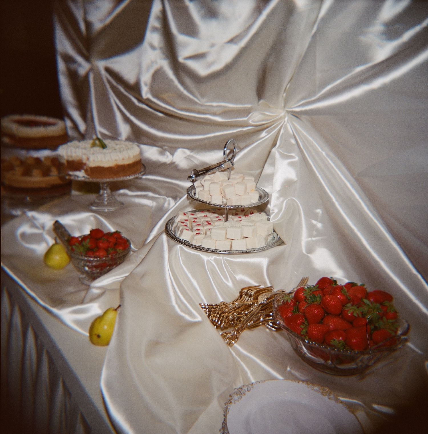 Elegant wedding dessert table with strawberries, cakes, and pears on white satin fabric.