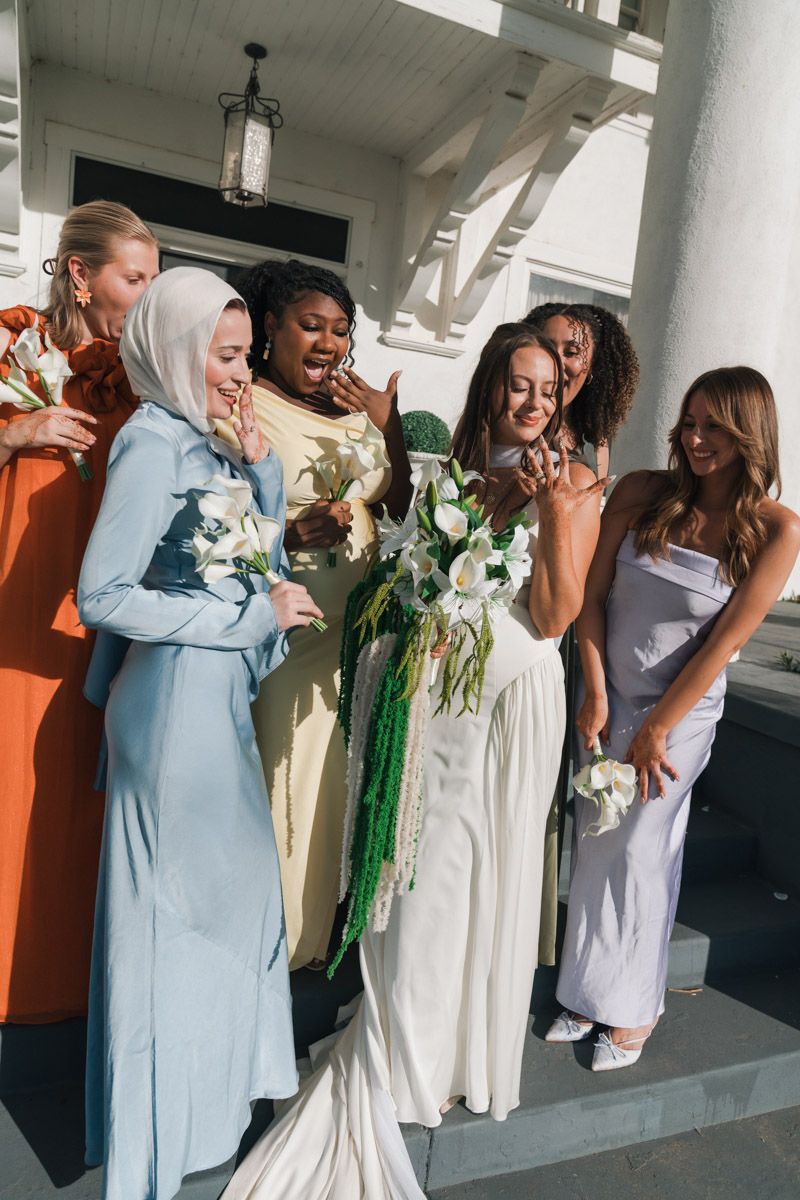 Group of bridesmaids in colorful dresses surround a bride in white holding a floral bouquet on a porch.