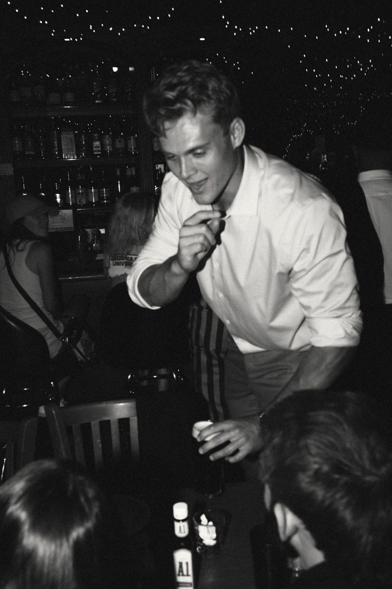 Black and white photo of a young man in a white shirt leaning forward at a bar, holding a drink.