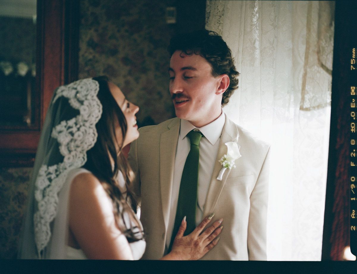 A bride and groom share a tender moment, smiling at each other in a warmly lit room on their wedding day on film.