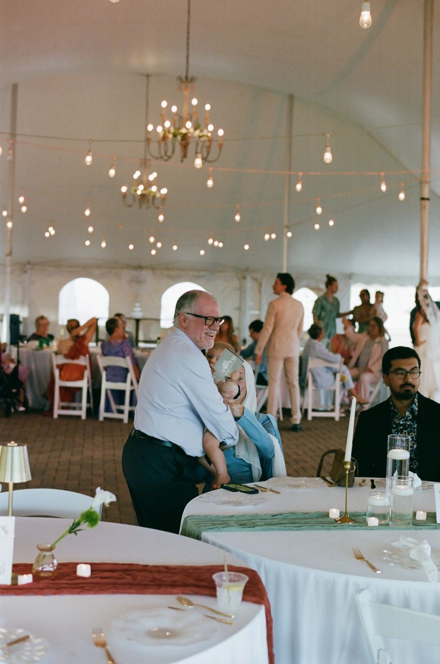 Wedding reception guests seated under chandelier-lit white tent with string lights overhead.