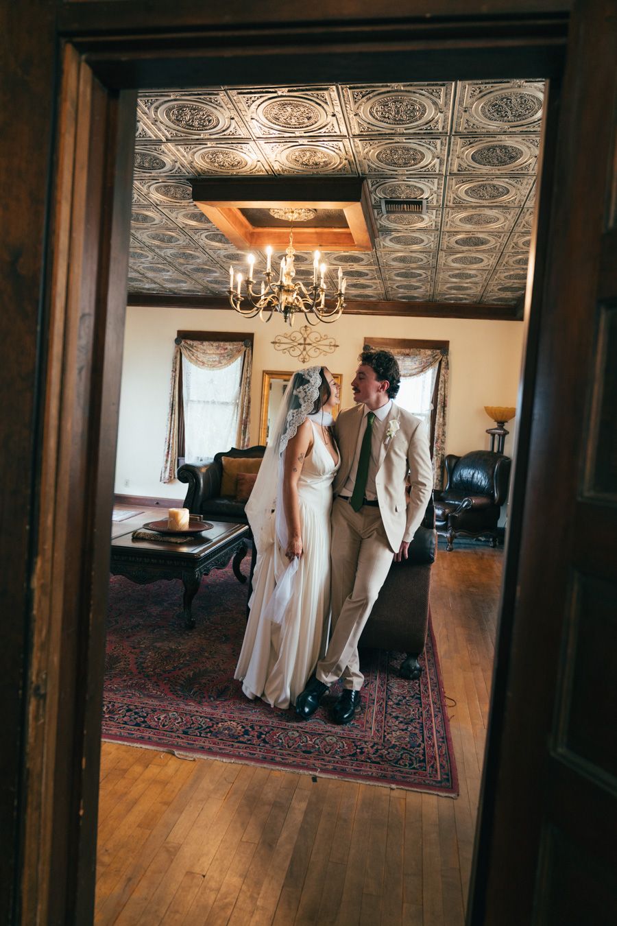 Bride and groom share intimate moment in vintage room with ornate tin ceiling and chandelier.