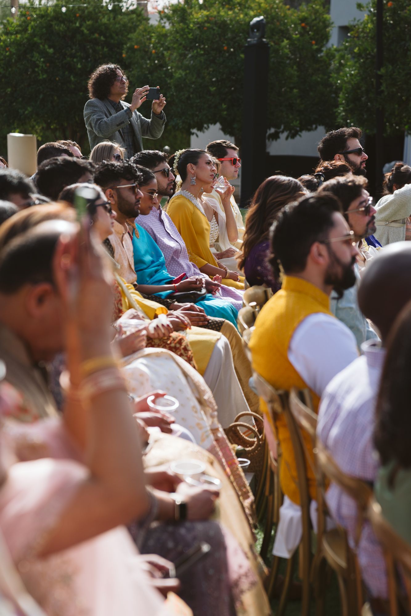 Wedding guests in colorful traditional Indian attire seated outdoors watching a sunny wedding ceremony.