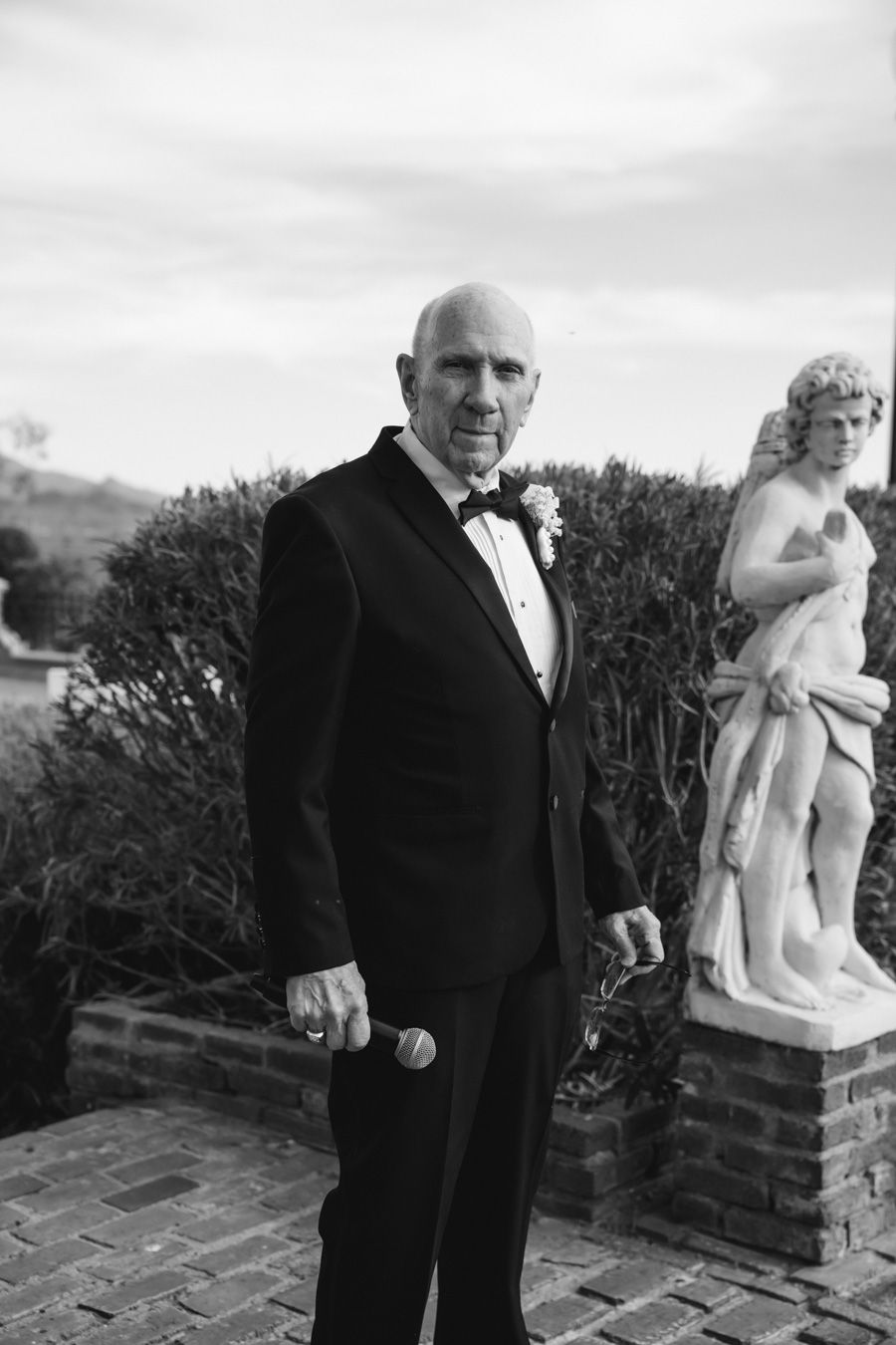 Officiant in tuxedo with boutonniere poses beside classical statue in black and white wedding photo at the end of the aisle.