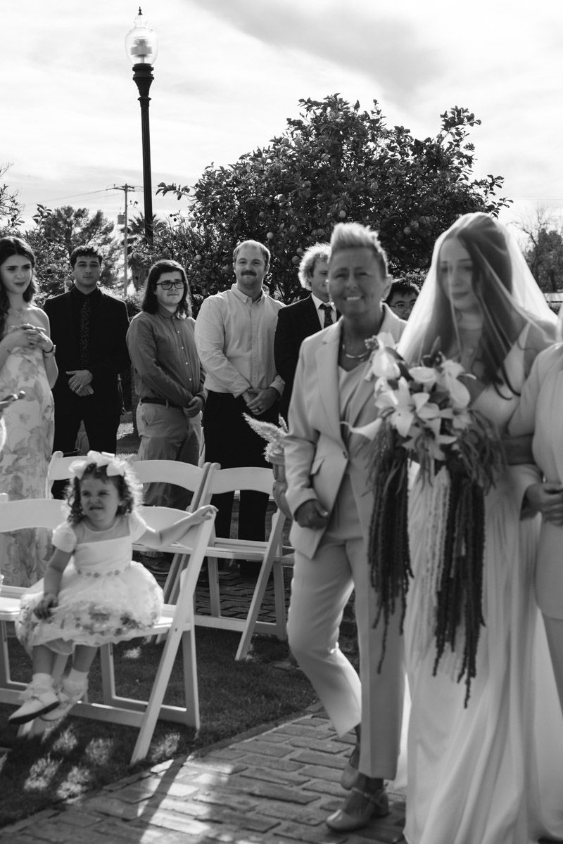 Black and white photo of a bride in veil holding bouquet walking down outdoor aisle with wedding guests watching.