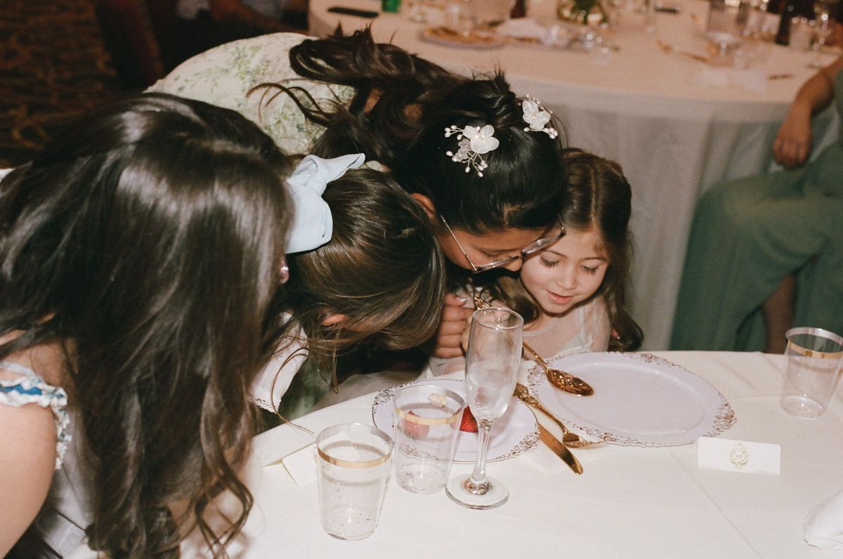 Children gathered around a wedding reception table, leaning in curiously to whisper to each other.