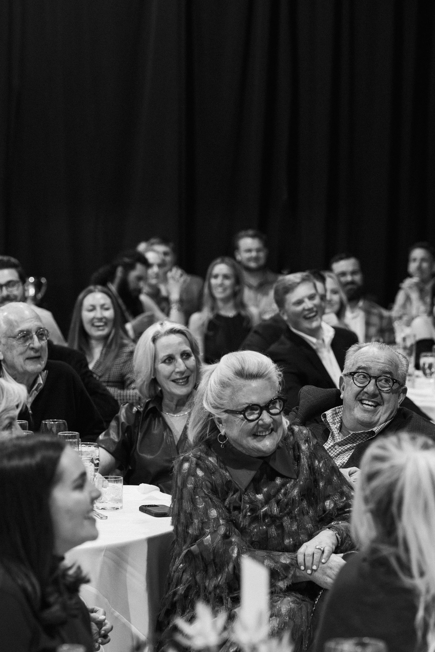 Black and white photo of smiling wedding guests seated at tables during a lively rehearsal celebration.