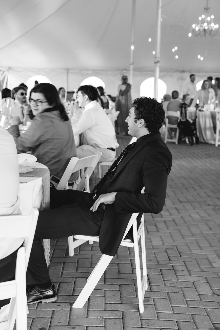 Wedding guest in black suit sits relaxed at wedding reception under white tent in black and white photo.